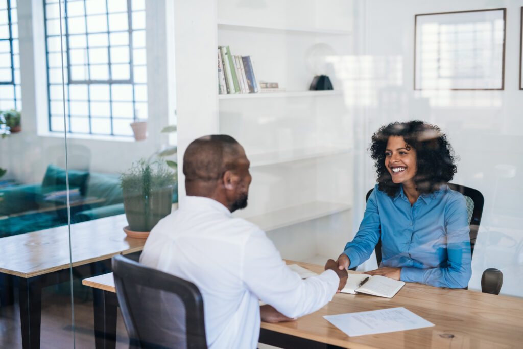 Smiling manager sitting at her desk inside of an office shaking hands with a potential new employee after an interview